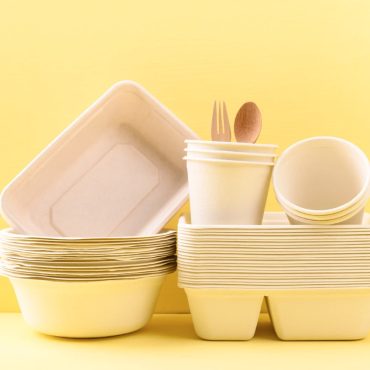 A stack of eco-friendly disposable tableware, including biodegradable cups, food containers, and wooden cutlery, arranged against a yellow background.