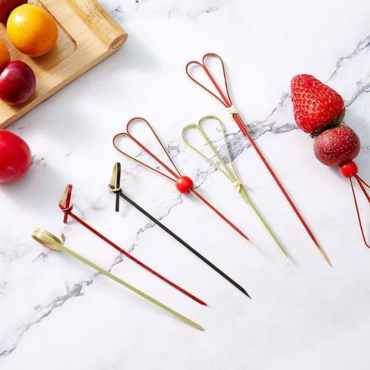 Colorful bamboo knot skewers in various colors used for serving fruits like strawberries, grapes, and oranges on a marble surface.